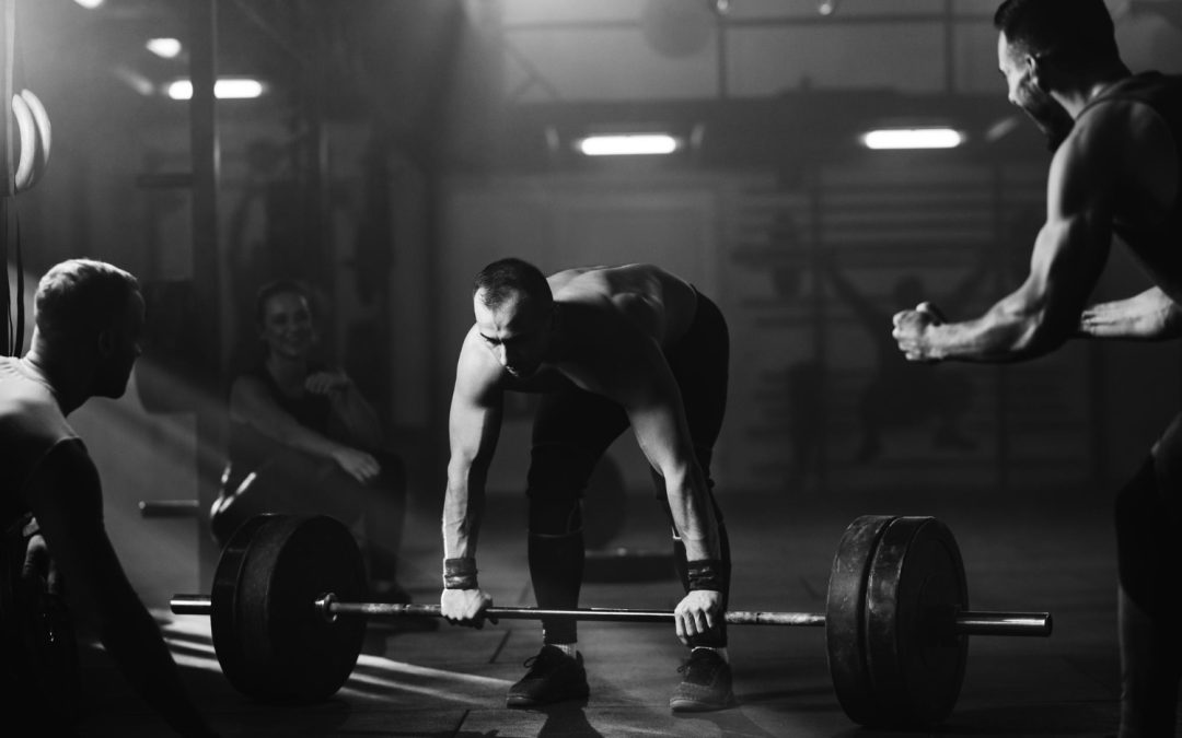 Man lifting a heavy barbell in a gym, surrounded by three cheering people, in dramatic lighting.
