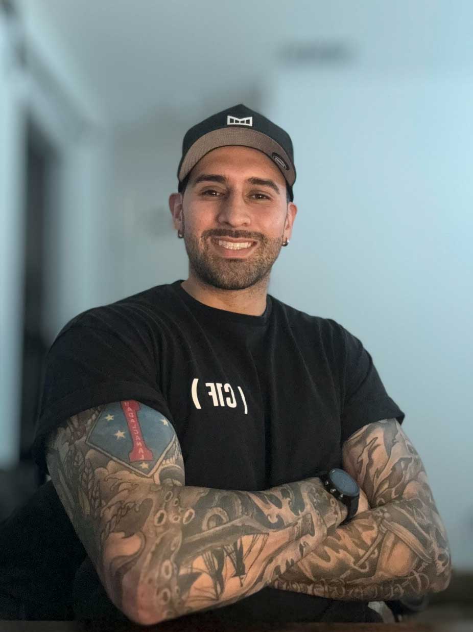 Smiling man with tattoos and a black cap sits with arms crossed, wearing a black t-shirt in a bright room.