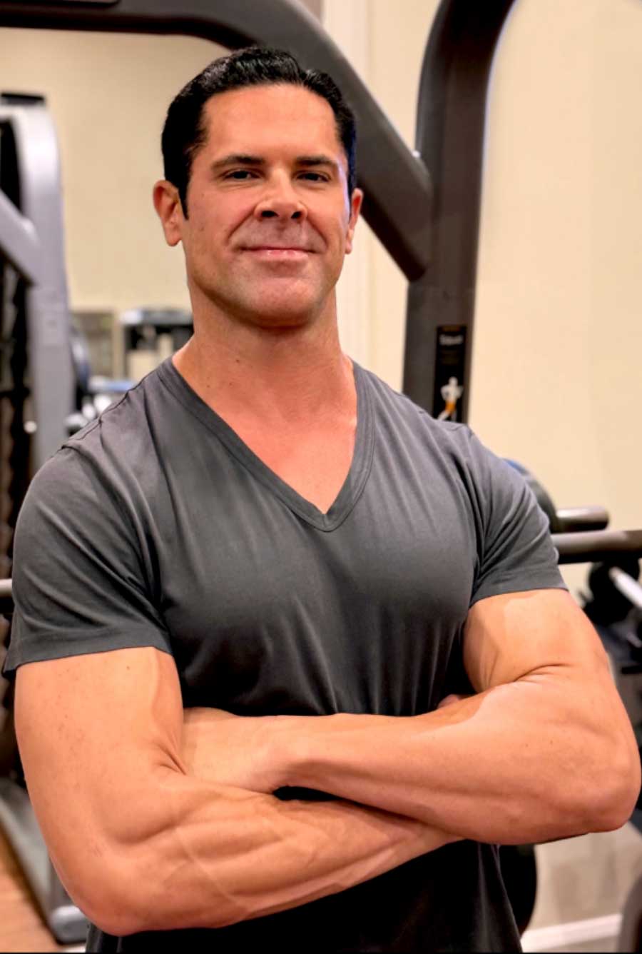 Muscular man with dark hair in a gray t-shirt stands with arms crossed in a gym.