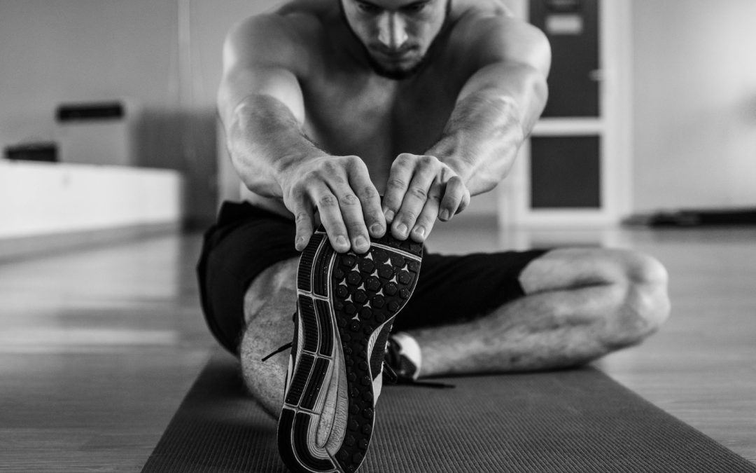 Shirtless man on a mat reaching forward to stretch his leg in a gym setting.