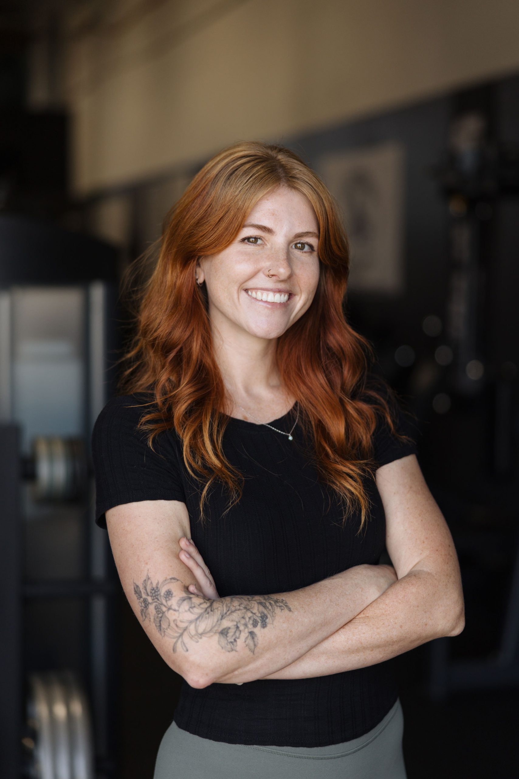 Smiling woman with long red hair and arm tattoos standing with arms crossed in a gym.