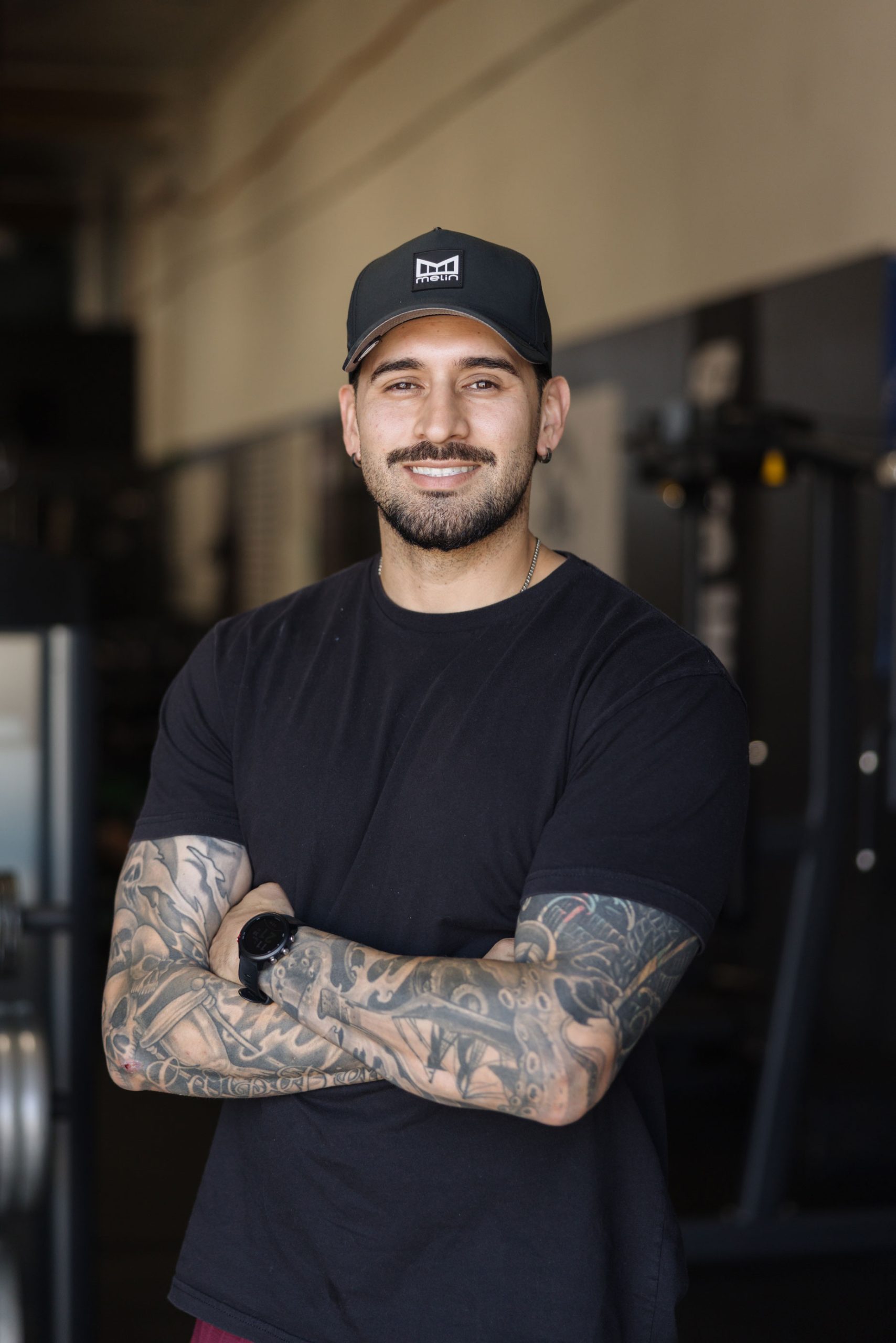 A smiling man with tattooed arms stands with his arms crossed in a gym.