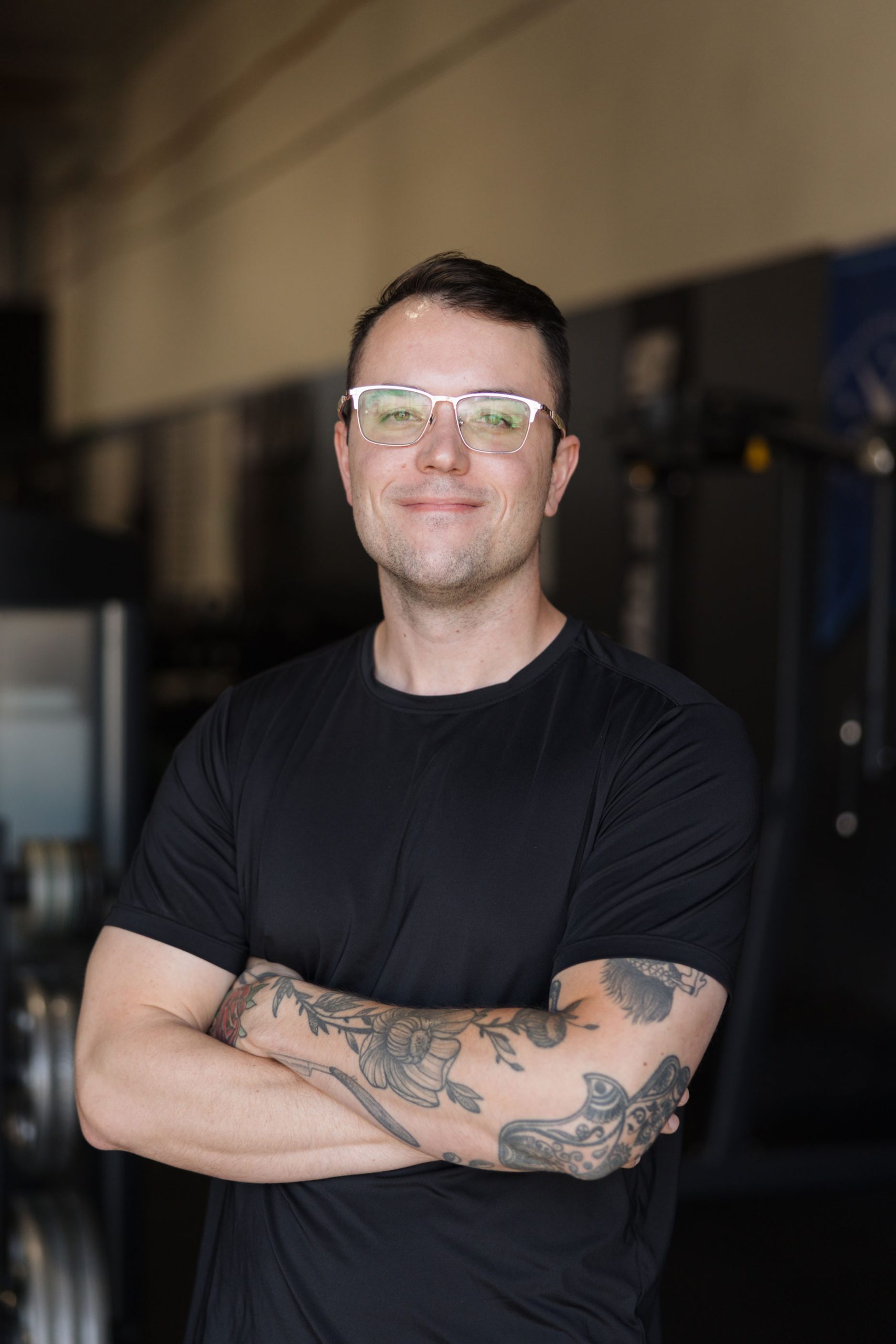 A man with glasses and arm tattoos standing with his arms crossed in a gym.