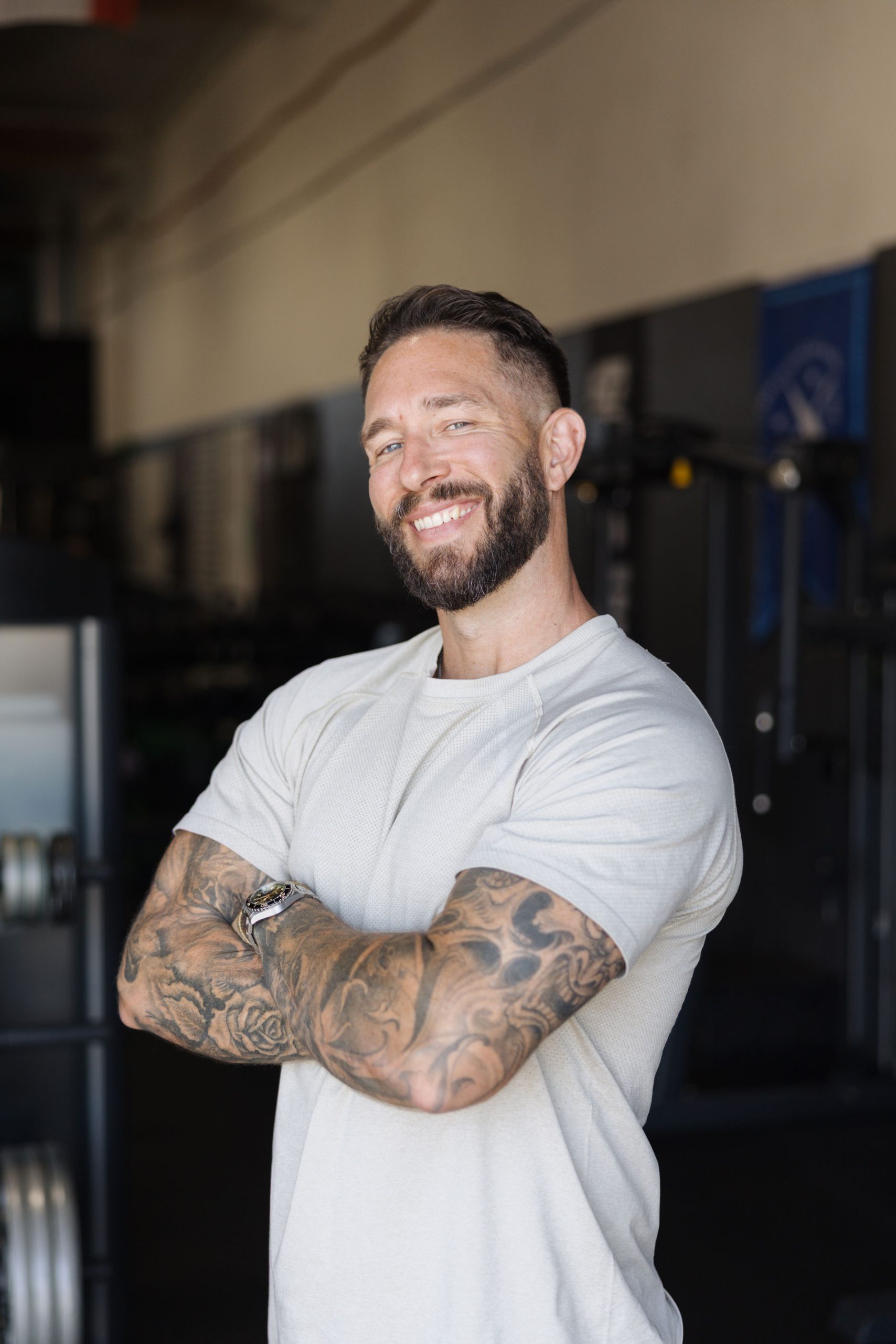 A smiling man with tattooed arms stands with his arms crossed in a gym.