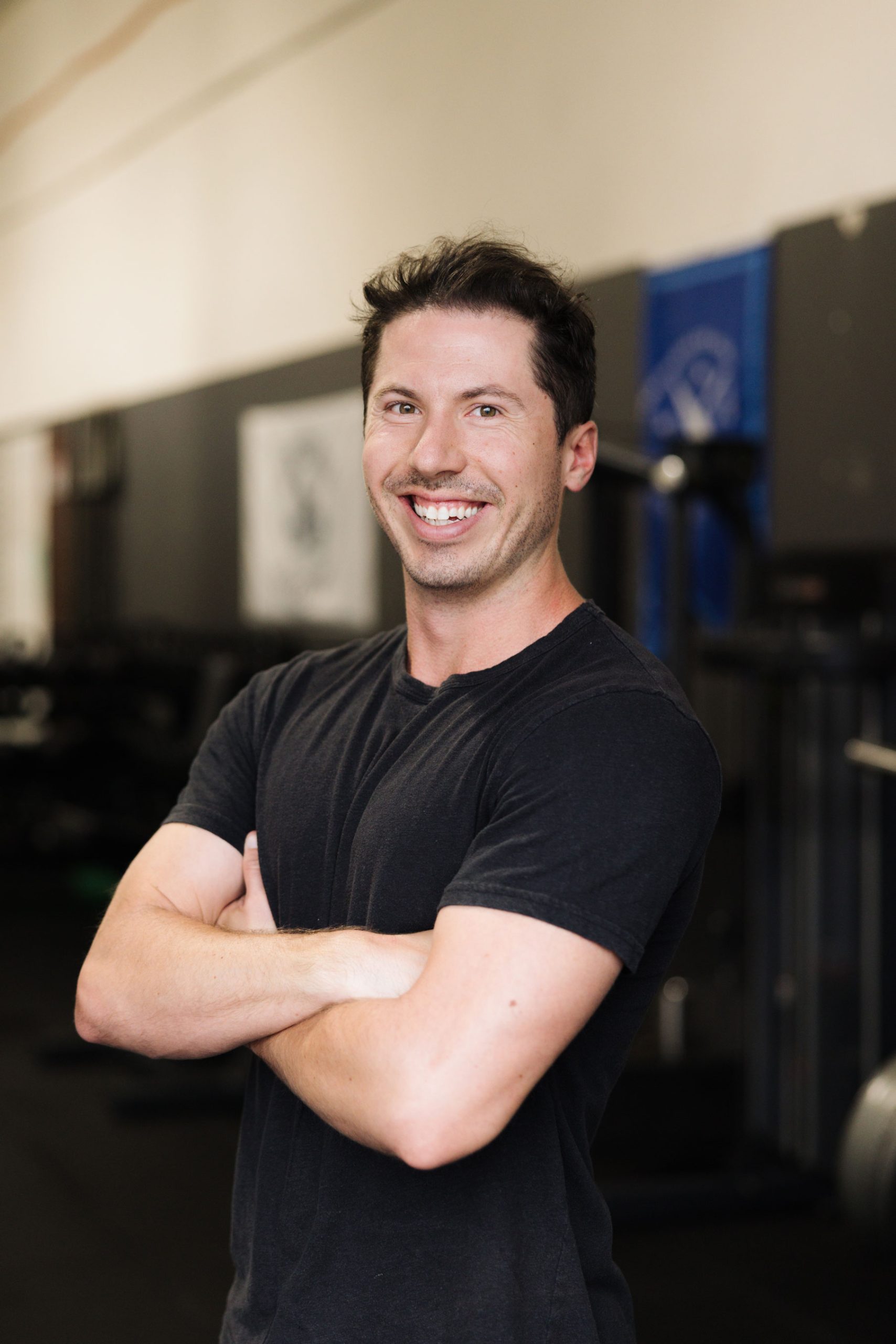 A smiling man in a black t-shirt standing with his arms crossed in a gym.