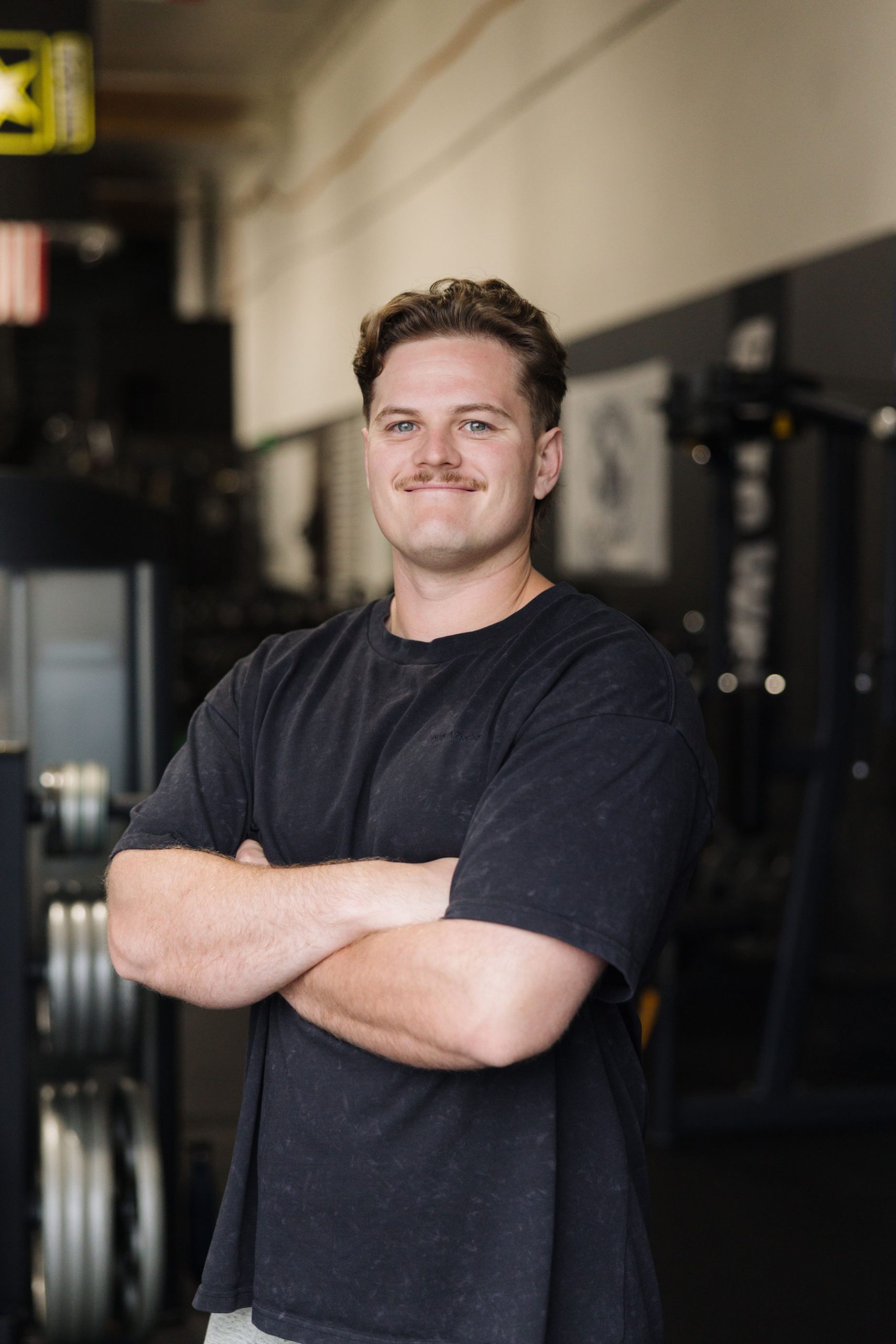 A man with a mustache stands in a gym with his arms crossed.